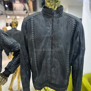 Pode incluir: Casaco preto com padrão de pele de cobra num manequim. O casaco tem gola alta e fecho de correr comprido. O manequim é dourado, com um espelho e outros manequins no fundo.