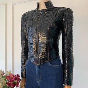 Peut inclure: Veste en cuir noir texturé, avec un motif serpent. Elle est dotée d'un col montant et d'une fermeture éclair intégrale. La veste a des manches longues et une coupe ajustée. Elle est portée avec un jean bleu. La veste est présentée sur un mannequin.