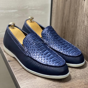 Può includere: Un paio di mocassini in pelle blu navy con un motivo a pelle di serpente sulla tomaia. Le scarpe hanno una suola bianca e sono esposte su una superficie di legno. Forme per scarpe all'interno.