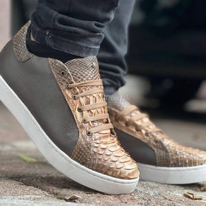 Könnte beinhalten: Braune und beige Ledersneaker mit weißen Sohlen. Die Sneaker haben ein Schlangenmuster auf Zehen und Ferse, mit braunen Lederpaneelen an den Seiten. Die Schnürsenkel sind beige, und die Schuhe werden mit einer schwarzen Hose getragen.