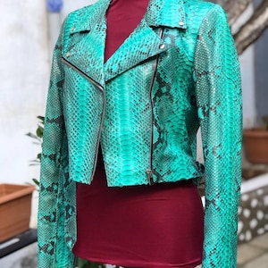 May include: A turquoise and black snakeskin-effect leather jacket in a classic biker style. The jacket has a notched collar, an asymmetrical zip, and long sleeves. The jacket is displayed on a mannequin with a burgundy top.