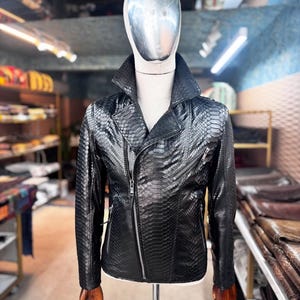 以下が含まれることがあります： マネキンに展示された、ヘビ革模様の黒い革のジャケット。 ジャケットは、非対称のジッパー開閉、広い襟、複数のジッパーポケットが特徴です。 ジャケットはミディアムサイズです。