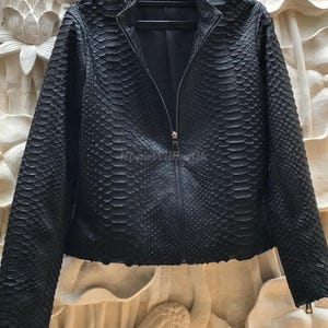 Può includere: Giacca in pelle nera con un motivo strutturato a forma di pelle di serpente. La giacca ha un collo alto, una cerniera a tutta lunghezza e cerniere sulle maniche. La giacca è appesa a una gruccia nera.
