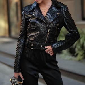 Könnte beinhalten: Schwarze Lederjacke mit Schlangenmuster, mit Reverskragen, diagonalem Reißverschluss und Gürtel. Die Jacke hat lange Ärmel und goldfarbene Hardware. Ein auffälliger und modischer Stil für verschiedene Anlässe.