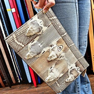 Può includere: Una pochette rettangolare con un motivo a pelle di serpente nei toni del beige, marrone e bianco. La pochette presenta decorazioni a testa di serpente tridimensionali. La borsa è tenuta su uno sfondo di cinture e jeans.