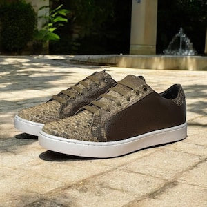 May include: A pair of brown and tan sneakers with a snakeskin pattern on the toe and side panels. The sneakers have brown laces, a brown leather heel, and a white sole. The shoes are displayed on a stone surface.