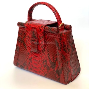 May include: A red handbag with a top handle and a flap closure. The bag features a snakeskin pattern in red and black. The handle is rounded and the bag has gold-coloured hardware.