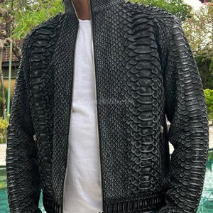 Pode incluir: Casaco de couro preto com padrão de pele de cobra. O casaco tem gola alta, fecho de correr e dois bolsos laterais. O casaco é usado sobre uma camiseta branca.