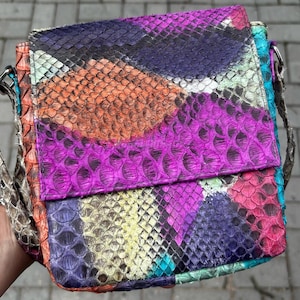 Può includere: Una colorata borsa a tracolla quadrata con patta. La borsa è realizzata in pelle di serpente con un design patchwork di viola, arancione, blu e rosa. La tracolla è di colore neutro.