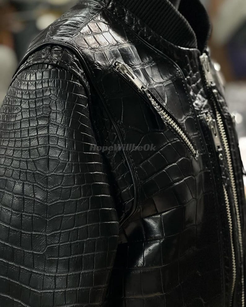 May include: Black leather jacket with a crocodile skin effect. The jacket features a ribbed collar and multiple silver zippers. The texture is detailed, suggesting a high-end garment.