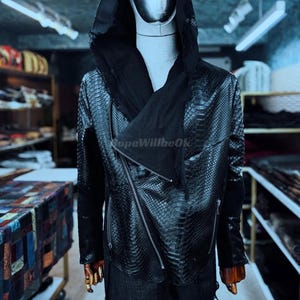 May include: Black faux leather jacket with a hood and asymmetrical zipper closure. The jacket has a snakeskin texture and a draped scarf detail. The text "HopeWillbeOk" is visible on the jacket.