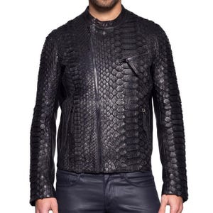 Può includere: Giacca in pelle nera con motivo a pelle di serpente. La giacca ha una chiusura con cerniera, una piccola tasca sul petto e un colletto alla coreana. Le maniche sono lunghe e aderenti.