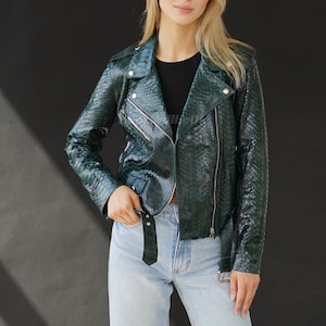 Puede incluir: Chaqueta de cuero verde oscuro con textura de piel de serpiente, de estilo motero clásico. La chaqueta presenta un cuello con solapas, cierre de cremallera asimétrico y cinturón en la cintura. Llevada sobre un top negro y vaqueros azules claros.