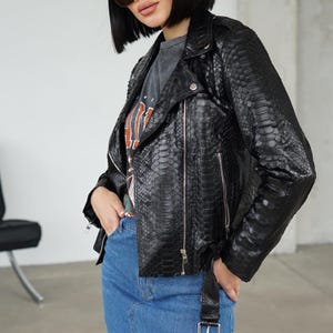 May include: A black leather jacket with a snakeskin texture. The jacket has a classic biker style with a notched collar, asymmetrical zipper, and a belt at the waist. The model is wearing a gray graphic tee and blue jeans.