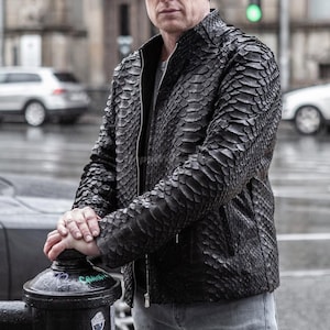 May include: A black leather jacket with a textured, scale-like pattern. The jacket has a zip closure and a stand-up collar. The person is wearing a grey shirt underneath. The background shows a city street with cars.