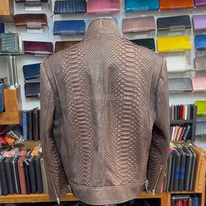 May include: A brown, snakeskin-effect leather jacket with a stand-up collar and zipped pockets. The jacket is displayed on a mannequin, showing its back. Shelves in the background display various wallets and bags.