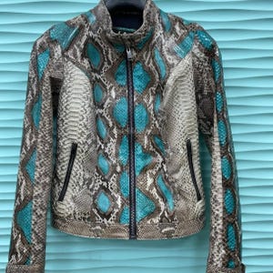 以下が含まれることがあります： ハイネックとフルレングスのジッパーが付いた、ヘビ革模様のジャケット。グレー、ブラウン、ティールブルーの色合いがミックスされています。ジッパーフロントと2つのフロントポケットを備えた、フィット感のあるシルエットです。