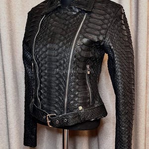 Op de afbeelding: Zwarte leren jas met een slangenpatroon. De jas heeft een reverskraag, een asymmetrische ritssluiting en een riem in de taille. Zilveren ritsen en hardware.