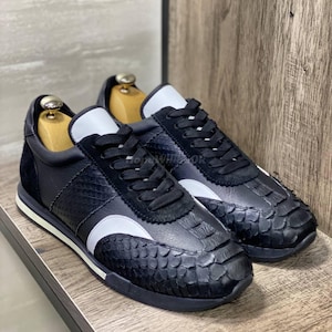 May include: A pair of black sneakers with white accents and a textured, scale-like pattern on the toe and heel. The shoes have black laces and a white stripe along the side. The sneakers are displayed on a wooden surface.