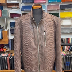 Pode incluir: Casaco de couro castanho com padrão de pele de cobra, exposto num manequim. O casaco tem fecho de correr frontal, um pequeno bolso no peito e gola alta. Várias carteiras de diferentes cores são visíveis.