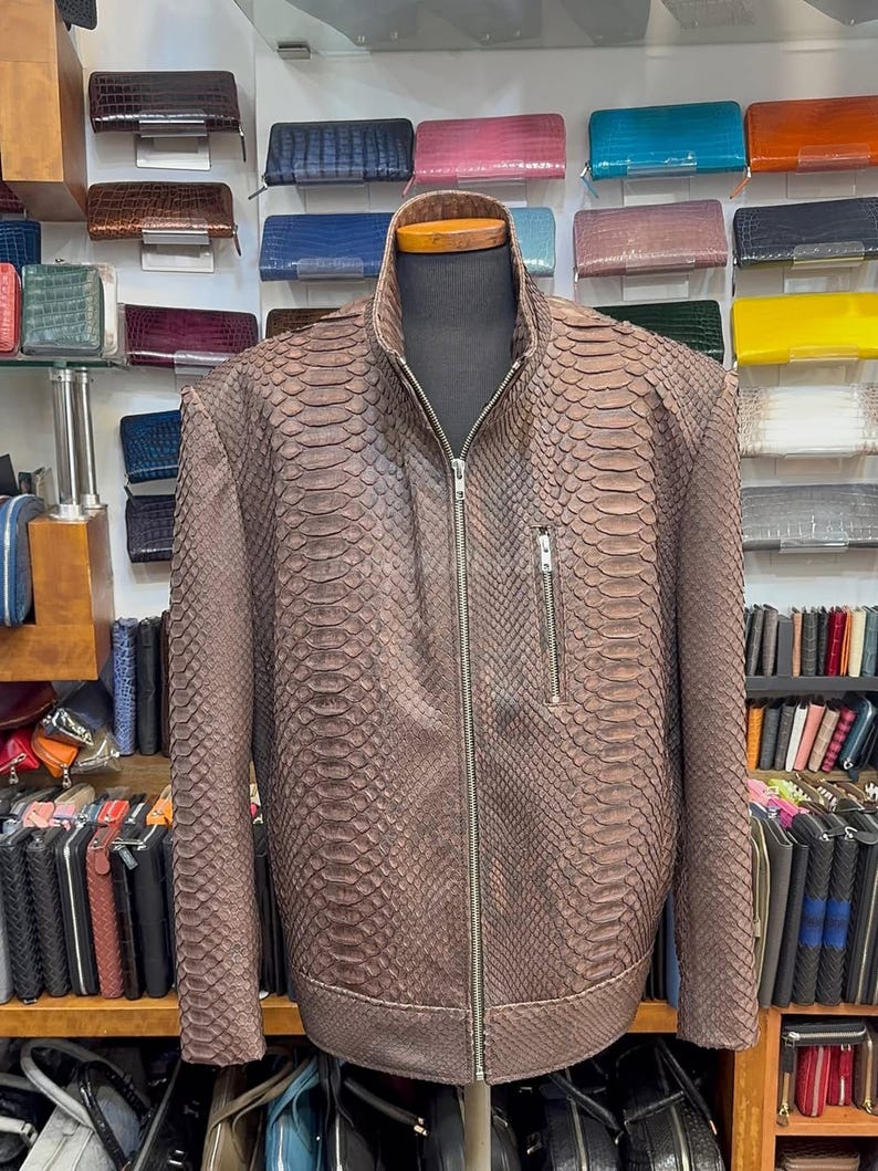 May include: Brown snakeskin-patterned leather jacket with a zip-up front and stand-up collar, displayed on a mannequin. A small chest pocket is visible. The jacket is surrounded by a variety of wallets in different colours.