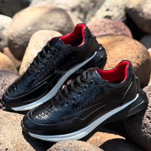 Puede incluir: Un par de zapatillas negras con un patrón texturizado y un interior rojo. Los zapatos tienen suelas blancas y cordones negros. Las zapatillas se exhiben sobre un lecho de rocas.