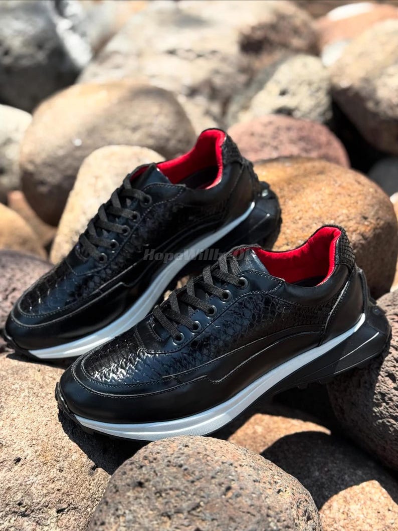 May include: A pair of black sneakers with a textured pattern and red interior. The shoes have white soles and black laces. The sneakers are displayed on a bed of rocks.