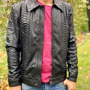 Può includere: Giacca in pelle nera con motivo a pelle di serpente. La giacca ha una chiusura a zip, un colletto e due tasche anteriori. La persona indossa una maglietta rosa e jeans blu.