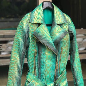 Może przedstawiać: Zielono-złota kurtka ze wzorem skóry węża, z paskiem w talii i złotymi zamkami błyskawicznymi. Kurtka ma klasyczny styl motocyklowy z wykładanym kołnierzem i długimi rękawami. Kurtka wisi na czarnym wieszaku.