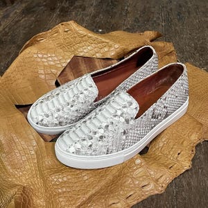 Puede incluir: Un par de zapatos blancos y grises sin cordones con un estampado de piel de serpiente. Los zapatos tienen suelas blancas y se exhiben sobre una superficie de cuero bronceado texturizado. Estilo casual.
