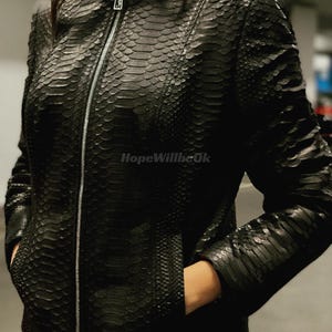 Può includere: Giacca in pelle nera con motivo a pelle di serpente, colletto alto e cerniera argentata. La giacca ha tasche ed è indossata da una persona. Il testo "HopeWillbeOk" è visibile.