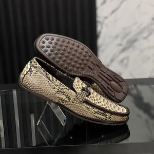 Könnte beinhalten: Schuhe mit Schlangenmuster, brauner Sohle und dekorativer Schnalle. Die Schuhe werden auf einem transparenten Ständer präsentiert, der das komplizierte Design und die Textur des Leders hervorhebt.