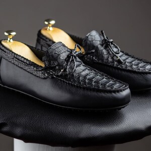 May include: A pair of black leather loafers with a snakeskin pattern on the vamp and heel. The shoes have a classic design with a rounded toe and a leather sole. The shoes are displayed on a black surface.
