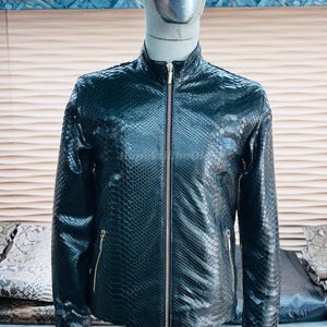May include: A black leather jacket with a snakeskin pattern is displayed on a mannequin. The jacket has a high collar, a full-length zipper, and two zippered pockets. The jacket is a dark color and has a shiny finish.