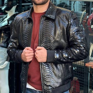 May include: A black leather jacket with a snakeskin pattern. The jacket has a zipper closure, ribbed cuffs, and a collar. The person is wearing a maroon shirt underneath. The jacket is a stylish outerwear piece.