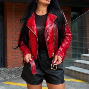 May include: A red snakeskin-effect jacket with a gold zip and buckle. The biker-style jacket features a notched collar and a waist belt. The model is wearing black shorts and a black top. The jacket is a bold fashion statement.