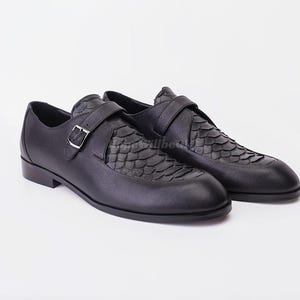 May include: A pair of black leather monk strap shoes. The shoes feature a textured vamp design and a silver buckle closure. The shoes have a low heel and a sleek, modern design, suitable for formal or semi-formal occasions.