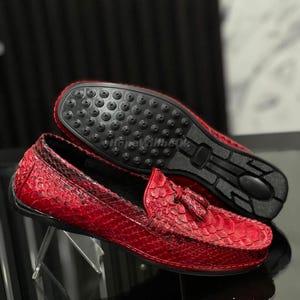 Pode incluir: Mocassins de couro vermelho com padrão de pele de cobra. Os sapatos têm detalhes de borlas e sola preta. Um sapato é exibido de cabeça para baixo, mostrando a sola.