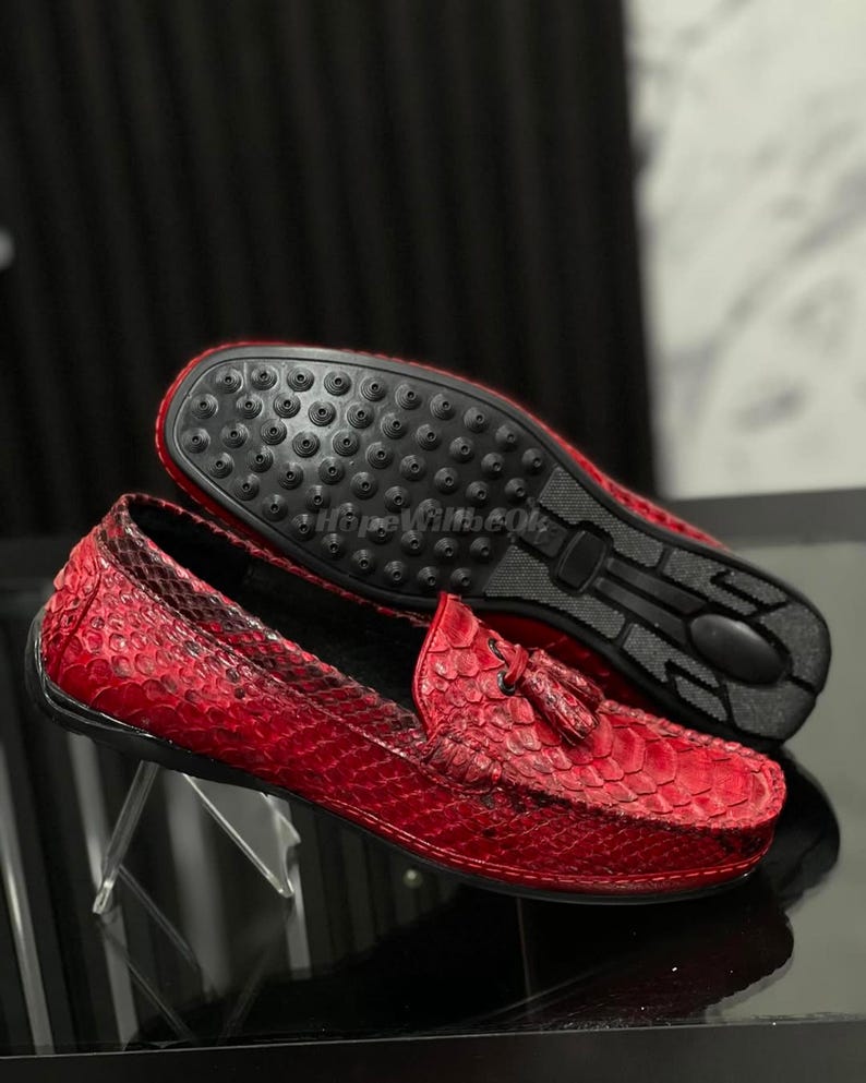 Op de afbeelding: Rode leren loafers met een slangenleerpatroon. De schoenen hebben kwastjes en een zwarte zool. E&eacute;n schoen wordt ondersteboven getoond, met de zool zichtbaar.