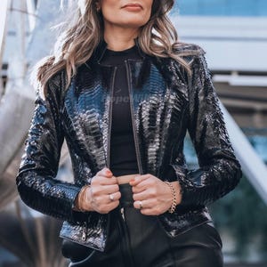 Op de afbeelding: Zwarte, getextureerde leren jas met ritssluiting. De jas heeft een slangenprint en een opstaande kraag. Het model draagt een zwarte top en een zwarte leren broek. De jas is het middelpunt van de afbeelding.