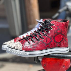 Puede incluir: Zapatillas altas con estampado de piel de serpiente roja y cordones negros. Las punteras son de color beige claro. Las zapatillas tienen suelas blancas y están sobre una superficie roja.