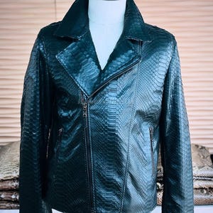 May include: Dark green leather jacket with a snakeskin pattern. The jacket features a classic asymmetrical zipper closure, a notched collar, and multiple zippered pockets. The jacket is displayed on a mannequin.