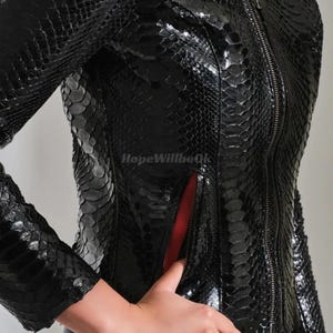 Może przedstawiać: Czarna sk&oacute;rzana kurtka o fakturze sk&oacute;ry węża. Kurtka ma zapięcie na zamek błyskawiczny i czerwoną podszewkę. Rękawy są długie, a kurtka dopasowana. Styl jest odważny i modny.