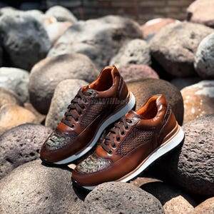 Könnte beinhalten: Ein Paar braune Ledersneaker mit strukturierten Einsätzen und reptilienartigen Akzenten an der Spitze. Die Schuhe haben weiße Sohlen und braune Schnürsenkel. Die Sneaker werden auf grauen Felsen präsentiert.