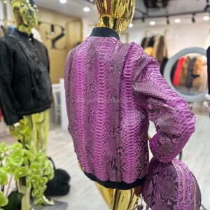 May include: A bright pink snakeskin-effect bomber jacket and matching small backpack. The jacket has a black ribbed collar and cuffs. The mannequin is gold-coloured. Other clothing items are visible in the background.