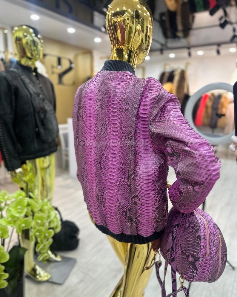 May include: A bright pink snakeskin-effect bomber jacket and matching small backpack. The jacket has a black ribbed collar and cuffs. The mannequin is gold-coloured. Other clothing items are visible in the background.