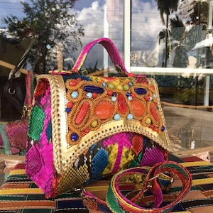 May include: A vibrant, multi-colored handbag with a snakeskin pattern. The bag features a gold-trimmed flap adorned with orange, red, and blue beads. It has a matching strap and handle, showcasing a bold and artistic design.