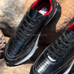 May include: Pair of black sneakers with a textured, reptile-like pattern. The shoes have black laces, a white sole, and a red interior. The sneakers are displayed on a rocky surface.