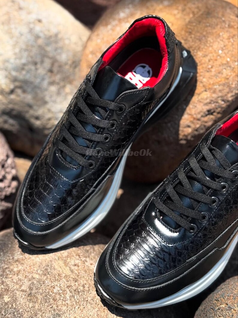 May include: Pair of black sneakers with a textured, reptile-like pattern. The shoes have black laces, a white sole, and a red interior. The sneakers are displayed on a rocky surface.
