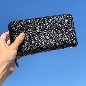 Puede incluir: Cartera negra con superficie texturizada y puntos blancos. La cartera tiene cierre de cremallera y se sostiene contra un cielo azul. El diseño de la cartera recuerda a la piel de raya.